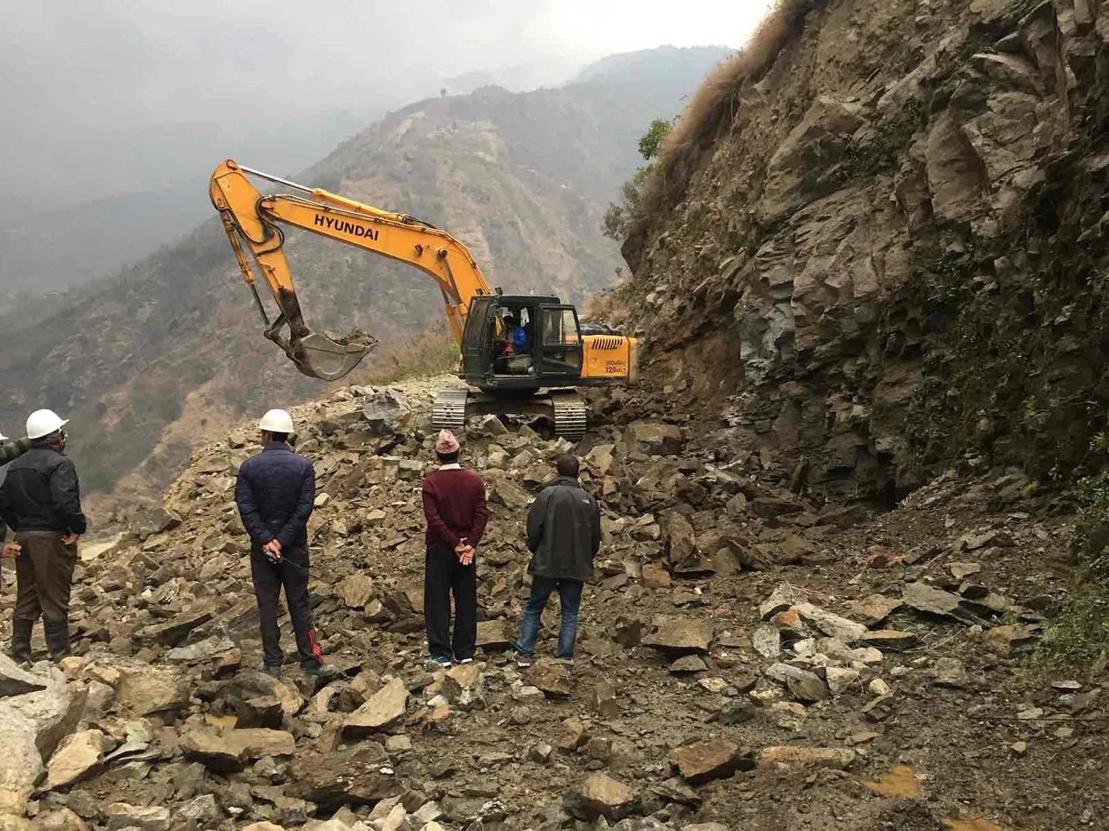 Excavator working a steep rock slope above the site, with three people surveying the ground